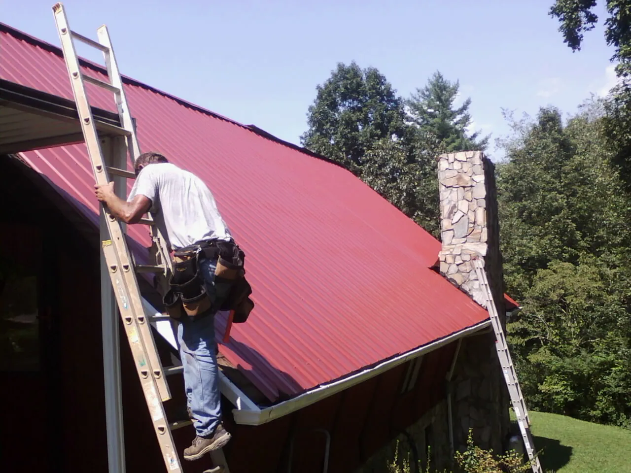 Roofer on a ladder beside a red metal roof and stone chimney — Soffit & Fascia Repair by Roofing Brothers in Donaldsonville